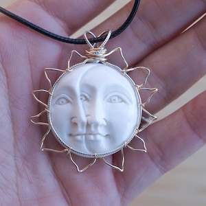 May include: A white pendant necklace with a carved sun and moon face design. The pendant is framed by a gold-coloured wire sunburst and a silver-coloured border. The necklace has a black cord.