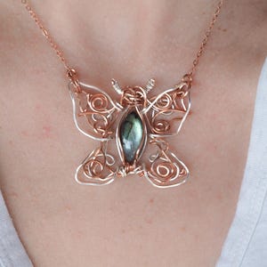 May include: A handcrafted butterfly pendant necklace. The pendant features a wire-wrapped design with copper and silver-toned metal. The butterfly has a central, oval-shaped, iridescent gemstone. The necklace chain is a delicate copper color.