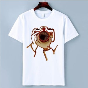 May include: White t-shirt featuring a coffee-themed design. The graphic depicts a coffee cup held like a camera, with the coffee forming the lens. The design is in shades of brown and red, creating a unique and artistic look.