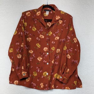 May include: A rust-colored button-down shirt with a floral pattern in shades of yellow, peach, and pink. The long-sleeved blouse has a collared neckline and is hanging on a black hanger. The shirt features a button-up front and buttoned cuffs.