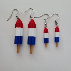 Pode incluir: Quatro pares de brincos em forma de picolé. Os brincos são vermelhos, brancos e azuis, com um palito de madeira. Os brincos estão em dois tamanhos, sendo que os maiores medem aproximadamente 5 cm de comprimento.