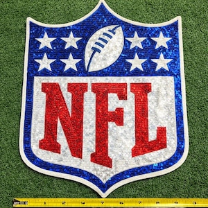 Puede incluir: Un logotipo de la NFL con lentejuelas en forma de escudo. El logotipo presenta un balón de fútbol blanco, estrellas blancas sobre un fondo azul y las letras "NFL" en rojo. El logotipo está delineado en blanco y azul, con un fondo verde.