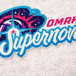 Könnte beinhalten: Ein paillettenbesetzter Aufnäher mit dem Schriftzug "Omaha Supernovas". Das Design zeigt einen Sternenstrahl und eine Farbpalette aus Blau, Weiß, Pink und Lila. Ideal für Kleidung oder Accessoires.