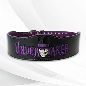May include: Black weightlifting belt with purple stitching and lining. The belt features the embroidered text "UNDERTAKER" in purple, along with an image of a man in a hat. The belt has a metal buckle.