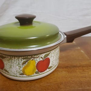 May include: A small, vintage saucepan with a green lid and brown handle. The pot features a white base with a fruit pattern of red apples and yellow pears. The lid has a brown knob.