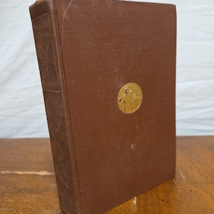 May include: A brown hardcover book with a gold emblem on the front cover. The book has a textured cover and a rectangular shape. The spine of the book has text. The book is leaning against a wooden surface.