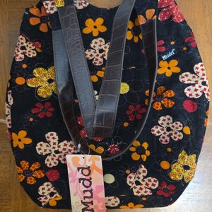 May include: A black corduroy tote bag featuring a colorful floral pattern with yellow, orange, and cream flowers. The bag has dark brown faux-leather straps with a crocodile-embossed texture and a Mudd brand tag attached to the front.