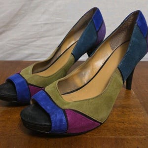 May include: A pair of high-heeled shoes with a color block design. The shoes feature a black platform sole, a blue toe strap, and panels of green, purple, and blue suede. The heels are black.