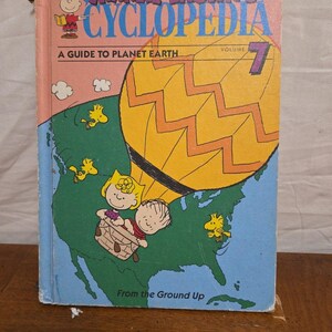 Könnte beinhalten: Ein Vintage-Buch "Charlie Brown's Cyclopedia", Band 7, mit einem Führer zum Planeten Erde. Das Cover zeigt einen gelben Heißluftballon über einer grünen Karte Nordamerikas, mit Snoopy und anderen Peanuts-Charakteren. Der Buchtitel ist in Blau und Rosa.