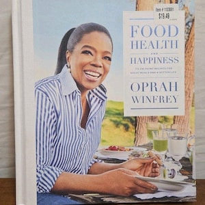 May include: A hardcover cookbook titled "Food Health and Happiness" by Oprah Winfrey. The cover features a photo of Oprah smiling, wearing a blue and white striped shirt. The book includes 115 recipes for great meals and a better life.