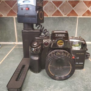 Könnte beinhalten: Schwarze Canon DL-9000 Kamera mit Blitzgerät. Die Kamera hat ein großes Objektiv und die Worte "OPTICAL LENS" sind sichtbar. Das Blitzgerät hat die Anzeigen "READY", "OFF" und "ON". Die Kamera steht auf einer gefliesten Oberfläche.