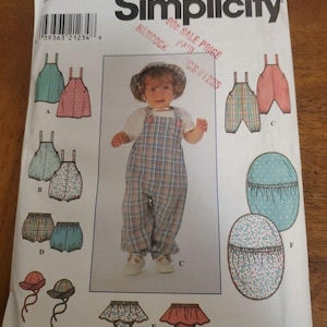 以下が含まれることがあります： Simplicityの型紙封筒、番号7996。オーバーオール、ブルマ、帽子などの乳幼児用衣類用。白い紙の封筒には、AからFまでのラベルが付いた様々なスタイルの衣類のイラストと、チェック柄のオーバーオールと帽子を着用した子供の中央写真が掲載されています。