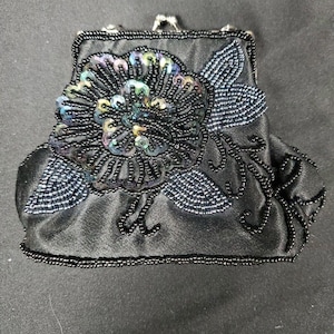 Puede incluir: Bolso de noche de satén negro con un diseño floral de cuentas. La flor presenta lentejuelas iridiscentes y cuentas negras. El bolso tiene un borde de cuentas y un cierre plateado.