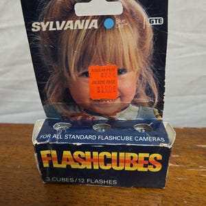 May include: Vintage Sylvania Flashcubes box with a child's image. The box is dark blue with the word "FLASHCUBES" in orange and yellow. It reads "3 CUBES/12 FLASHES" and "FOR ALL STANDARD FLASHCUBE CAMERAS".