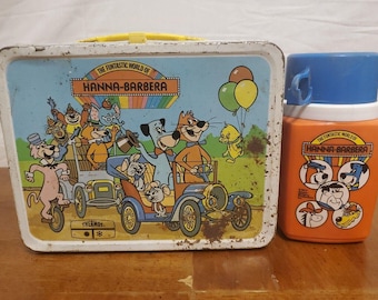 May include: A vintage Hanna-Barbera lunchbox and thermos set. The lunchbox has a colourful illustration of cartoon characters in a car, with the text "THE FUNTASTIC WORLD OF HANNA-BARBERA". The orange thermos has a blue lid and matching graphics.
