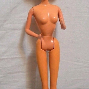 May include: A plastic doll with light peach skin tone, blue eyes, and short, curly blonde hair. The doll has a slender build, articulated arms and legs, and a smiling facial expression. It is standing upright with its arms bent at the elbows.