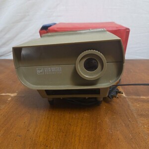 May include: A beige GAF View-Master projector featuring a circular lens on the right side and the brand logo with text printed on the front. The device includes a black power cord and is positioned in front of a red storage case.
