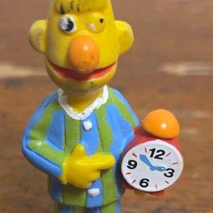 May include: A yellow figurine of a character with a tall head, wearing blue and green striped pajamas and pink slippers. The character has a round orange nose and is holding a small red alarm clock with a white face, black numbers, and blue hands.