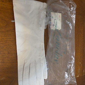 May include: A pair of long, off-white leather gloves, one laid out and the other still in its original plastic packaging. The glove is smooth and has a classic design. The packaging has a tag attached.