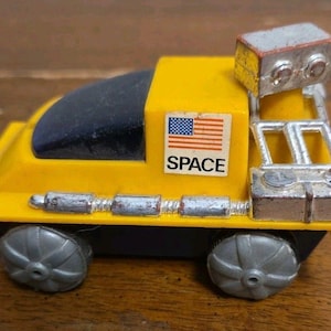 May include: A yellow toy space vehicle with a black windshield, silver wheels, and a silver rear structure. A sticker on the side displays the United States flag and the word SPACE in black capital letters. The toy features a metallic silver component.