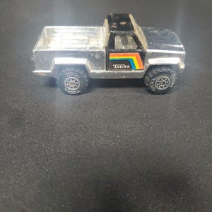 May include: A silver-colored toy pickup truck with a black cab section. The side of the truck features a graphic with a rainbow stripe and the word Tonka printed in white. The vehicle has black wheels with textured tires and a metallic silver truck bed.