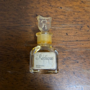 May include: A small, vintage perfume bottle with a clear glass body and a yellow stopper. The stopper is shaped like a stylized letter "R". The bottle has a label with the word "Réplique" in cursive script. The bottle is on a wooden surface.