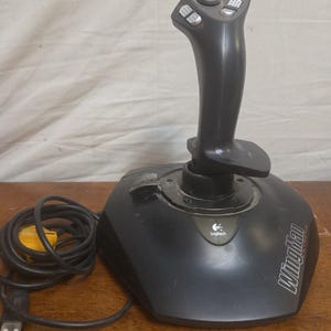 Puede incluir: Un joystick Logitech WingMan Extreme negro con una empuñadura vertical con varios botones, un interruptor de sombrero montado en la parte superior y una base con palanca de aceleración. Incluye un cable USB negro con conector amarillo.