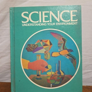 Puede incluir: Un libro de texto de ciencias turquesa titulado "SCIENCE UNDERSTANDING YOUR ENVIRONMENT". La portada presenta una ilustración circular con un pájaro en vuelo, una mano vertiendo líquido en un vaso de precipitados y formas geométricas. El libro está sobre una superficie de madera.