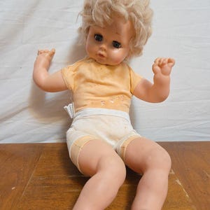 May include: A vintage baby doll with blonde, tousled hair, wearing a yellow short-sleeved shirt and white shorts. The doll has a sweet expression and is posed with arms raised. The doll is made of plastic and is sitting on a wooden surface.