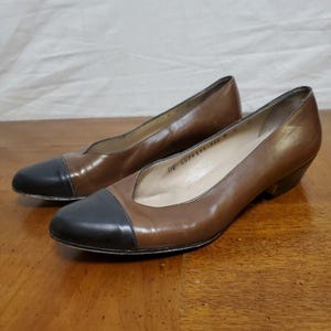 Könnte beinhalten: Ein Paar braune Lederpumps mit schwarzen Kappen. Die Schuhe haben einen niedrigen Absatz und ein klassisches Design, das für verschiedene Anlässe geeignet ist. Die Schuhe werden auf einer Holzoberfläche präsentiert.