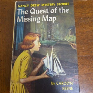 May include: A vintage hardcover book titled "Nancy Drew Mystery Stories: The Quest of the Missing Map" by Carolyn Keene. The cover art features a woman in a yellow dress holding a model sailboat, with a ship and a man in the background.