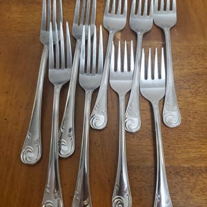 Puede incluir: Un conjunto de tenedores plateados con un patrón decorativo de remolino en los mangos. Los tenedores están dispuestos sobre una superficie de madera, mostrando su acabado pulido y su elegante diseño. Las púas están espaciadas uniformemente.