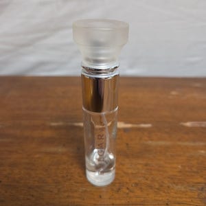 May include: A clear glass perfume bottle with a silver base and a frosted cap. The bottle has the word "BVLGARI" printed vertically on the side. The bottle is sitting on a wooden surface.