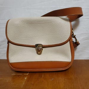 Pode incluir: Bolsa transversal de couro creme com acabamento e alça de couro marrom. A bolsa possui fecho de aba com fecho de metal dourado. A bolsa está em uma superfície de madeira.