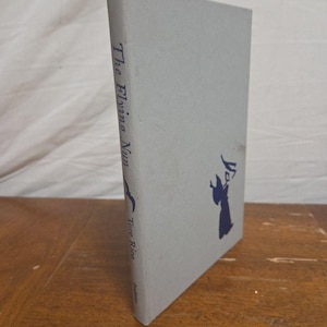 May include: A vintage book titled "The Flying Nun" with a light gray cover. The title and author's name are printed vertically in dark blue. A dark blue silhouette of a nun is on the cover.