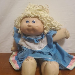 May include: A vintage doll with a soft, fabric body and a vinyl head. The doll has blonde yarn hair, blue eyes, and a rosy complexion. It wears a blue dress with a white and pink trim.