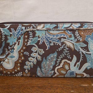 May include: A rectangular brown wallet with a floral pattern in shades of blue, teal, and tan. The wallet has a zipper closure and a small fabric loop on the side. The pattern features stylized leaves and floral elements.