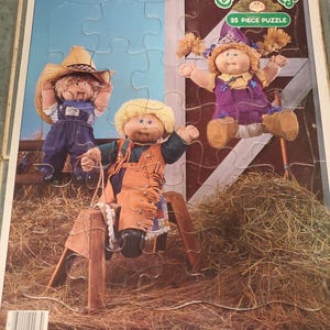 May include: A 25-piece Cabbage Patch Kids jigsaw puzzle featuring three dolls in a barn setting with hay. One doll wears a straw hat and overalls, another sits on a wooden sawhorse, and the third wears a purple outfit. The MB logo is visible.