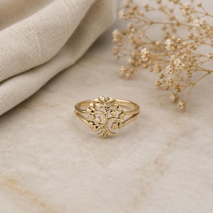 May include: A gold-toned ring featuring a circular centerpiece with a detailed tree of life design. The band is split into two thin, polished gold-toned segments that connect to the central tree motif, creating a cohesive and symmetrical appearance.
