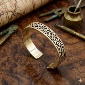 May include: A gold-colored cuff bracelet with an intricate Celtic knot design. The open-ended bracelet is displayed on a dark wooden surface, with antique tools and a map in the background. The bracelet's surface has a polished finish.