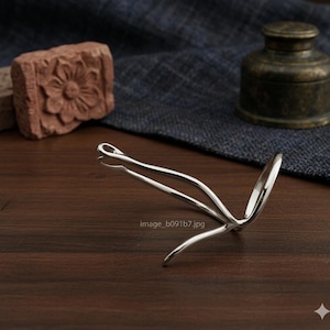 Puede incluir: Un accesorio para el cabello plateado con un diseño curvo único. El accesorio está sobre una superficie de madera oscura. En el fondo, una piedra tallada y un pequeño recipiente de metal.