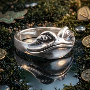 Puede incluir: Un anillo de plata con diseño de rana. El anillo presenta una cabeza de rana detallada con ojos visibles y piel texturizada. Se encuentra sobre un fondo de musgo verde y pequeñas monedas, creando un entorno natural.