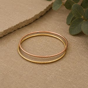 Op de afbeelding: Twee gouden armbanden, één glad en één met een textuurpatroon, worden getoond op een neutraal oppervlak. De bovenste armband heeft een roségoudkleur, terwijl de onderste een heldere goudkleur heeft. De sieraden zijn eenvoudig en elegant.