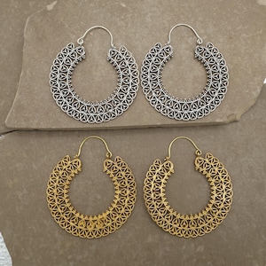 Op de afbeelding: Twee paar oorringen. Eén paar is zilverkleurig en het andere goudkleurig. Beide paren hebben een gedetailleerd, open ontwerp met een halvemaanvorm. De oorbellen worden gepresenteerd op een neutraal, steenachtig oppervlak.