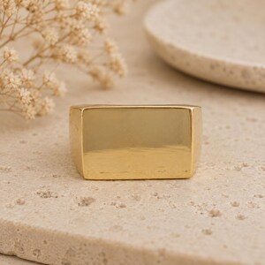 May include: A gold-colored, rectangular ring with rounded edges. The ring is a solid, polished metal with a smooth surface. The ring is set on a textured, light-colored surface.
