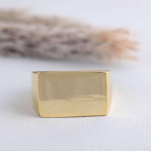 May include: A gold-colored signet ring with a rectangular face. The ring has a polished finish and a simple, classic design. The ring is likely made of a precious metal.