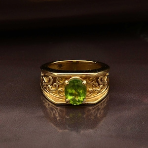 May include: A gold ring with an oval, light green gemstone at the center. The ring has an ornate, engraved design around the band. The ring is set against a dark background.