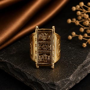 Könnte beinhalten: Ein goldfarbener Ring mit aufwendigen geometrischen Mustern. Der Ring hat ein rechteckiges Mittelstück mit detaillierten Designs und liegt auf einer dunklen, strukturierten Oberfläche. Das Design des Rings deutet auf einen Vintage- oder Antik-Stil hin, für Schmuckliebhaber.