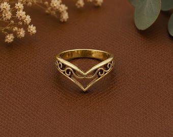May include: A gold-colored ring with a V-shaped design. The ring features an open center and decorative scrollwork along the top edges. The ring is set against a brown background, with sprigs of dried flowers and greenery in the background.