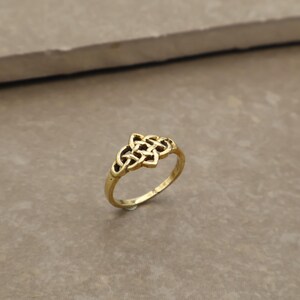 May include: A gold ring with a Celtic knot design. The ring has a slender band and a detailed knot centerpiece. The ring is shown on a neutral surface, emphasizing its intricate design and warm colour. The ring is likely made of a gold alloy.